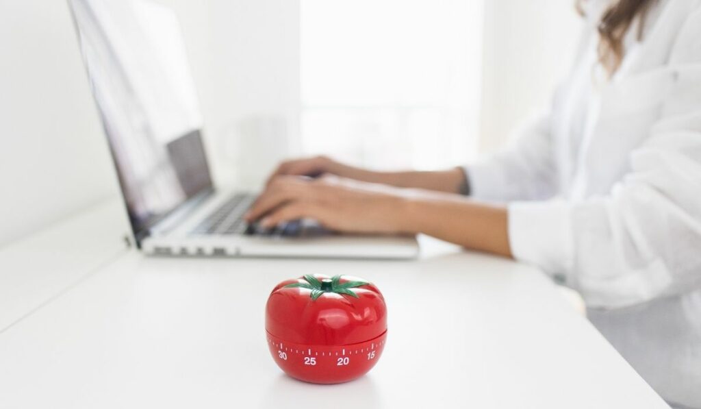 A tomato timer is placed on white desk while a woman is typing on her laptop