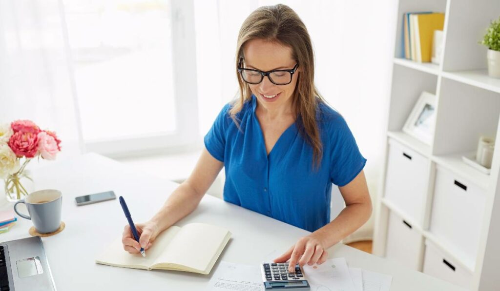 A middle-aged woman with glasses sitting at a desk with a calculator, book and laptop