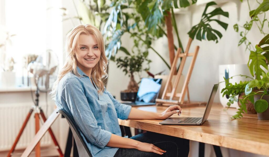 A smiling eco-conscious entrepreneur is sitting on a chair at a desk with a laptop.