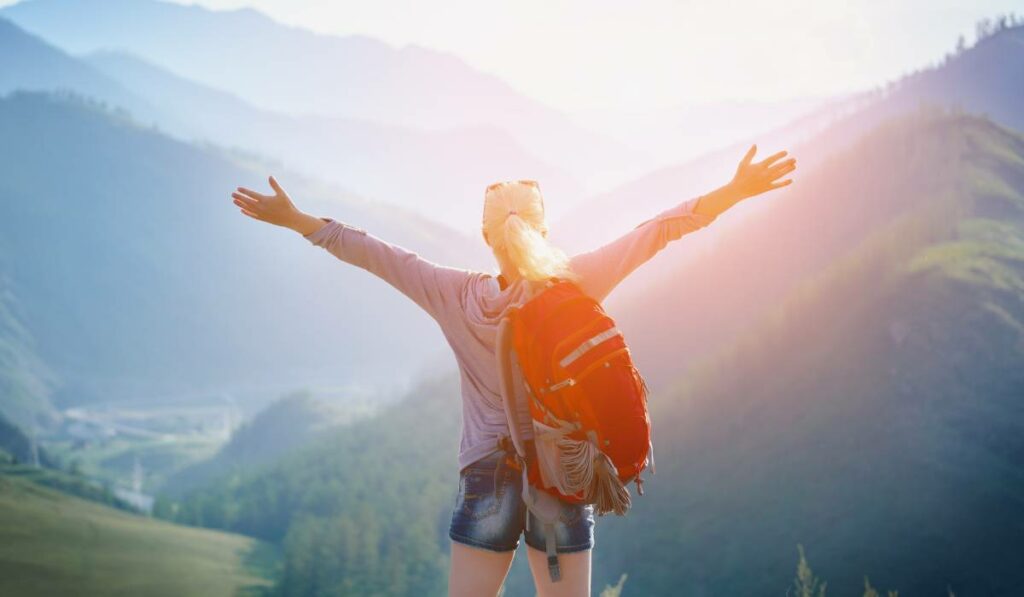 A woman with backpack standing on top of mountain with arms outstretched is exploring eco-tourism.