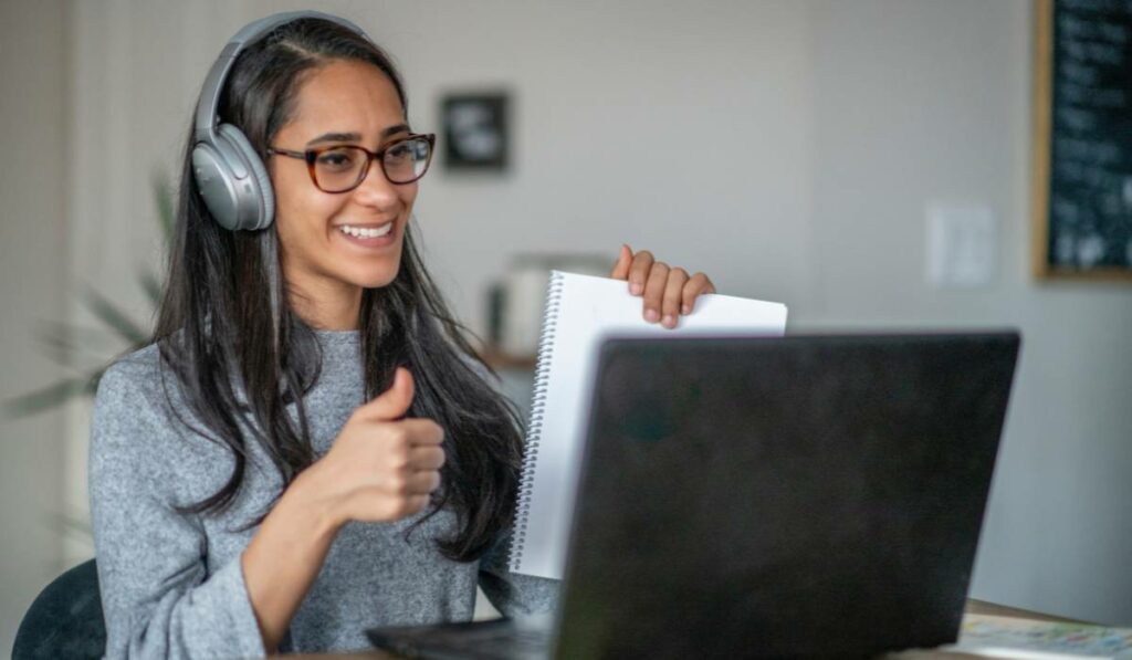 A woman smiling and giving a thumb up. She is wearing headphones, glasses and sitting in front of a laptop providing virtual assistance as a part of her side hustle.