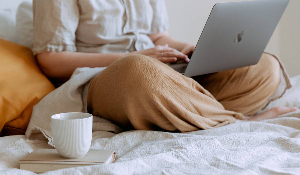A woman starting a side hustle, is sitting on a bed with a laptop and a coffee cup by her side.