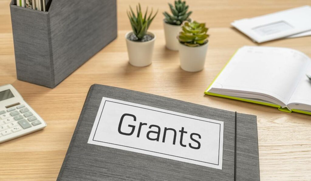 "Grants" written on a folder on a desk with a calculator, notepad, envelopes and plants on the background.