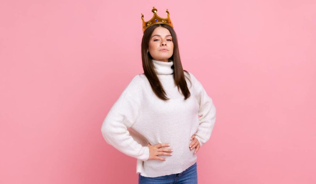A confident female entrepreneur wearing a crown and standing with arms on her waist against a pink background.